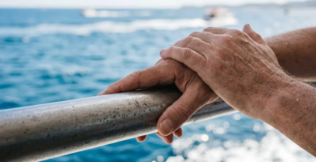 Mani di una persona appoggiate sulla ringhiera metallica di un traghetto, mare Mediterraneo sfocato sullo sfondo con riflessi di luce sull'acqua